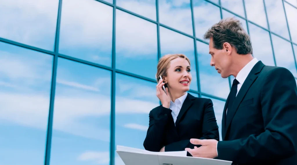 Ejecutivos de una empresa tecnológica conversan frente a un moderno edificio; la mujer utiliza un teléfono móvil, símbolo de comunicación y conectividad.